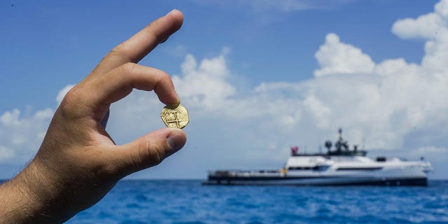 アレンの探査船が遠くから見えるバハマで発見された金貨を手にする探検家。
